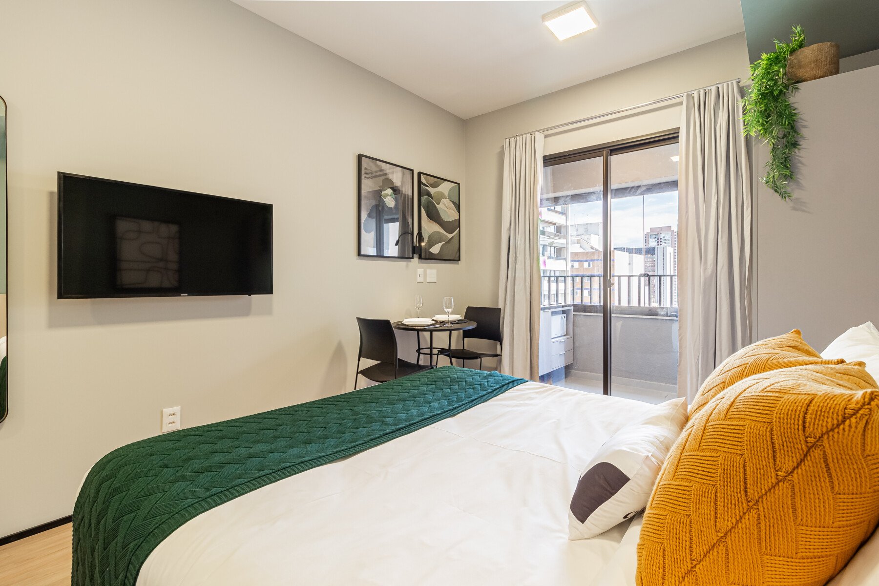 Quarto moderno e aconchegante com cama confortável, TV, mesa para refeições e varanda, em tons neutros e detalhes coloridos.