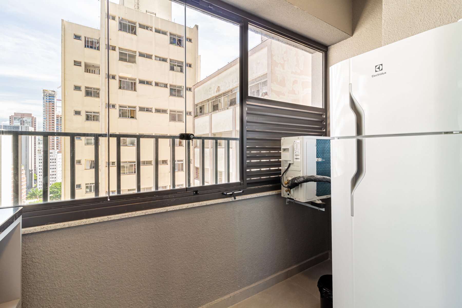 Imagem de uma varanda de apartamento, mostrando uma geladeira moderna com vista para a cidade, destacando o espaço urbanizado e a iluminação natural.