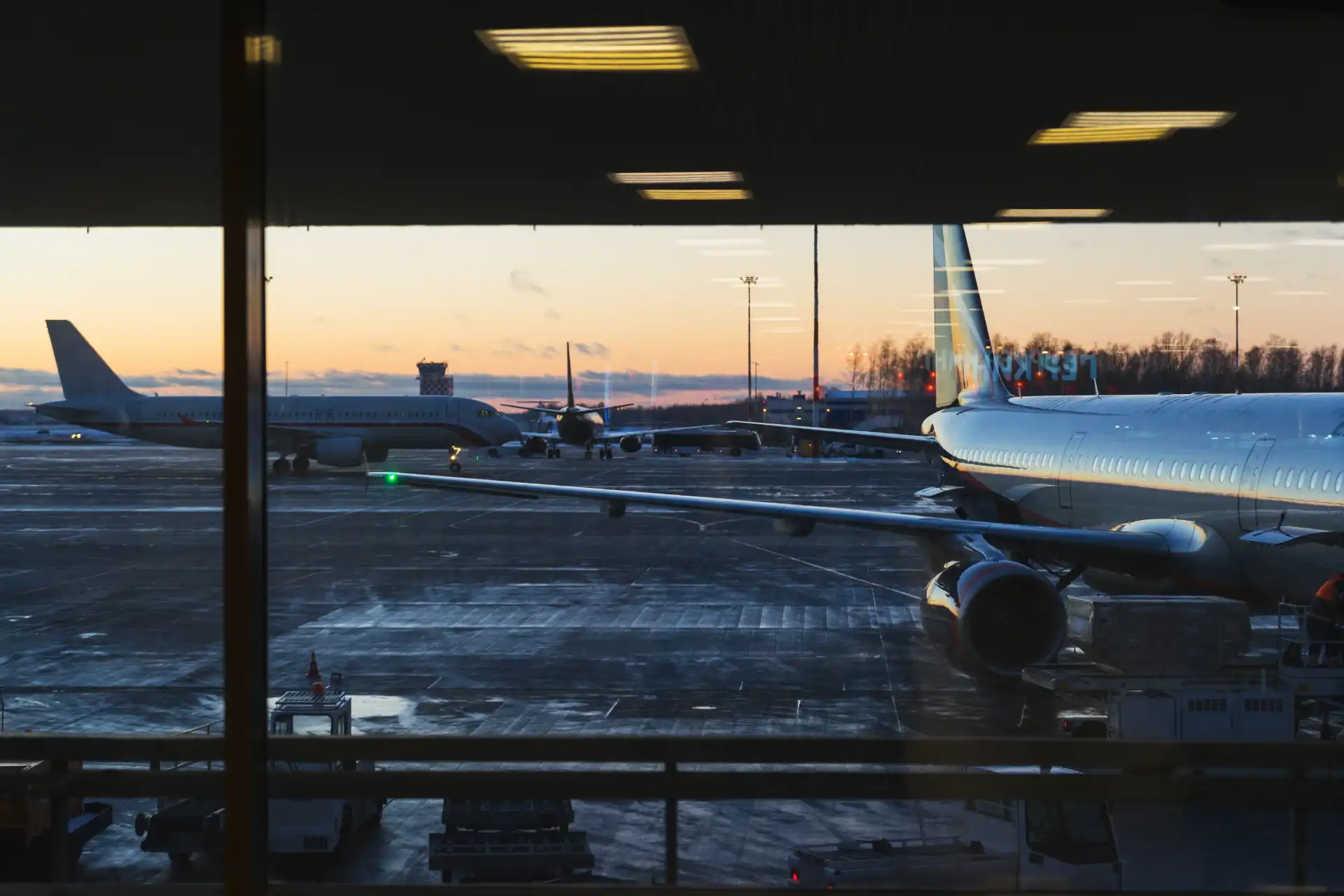 Imagem de aeroporto mostrando aviões estacionados ao entardecer, com luzes acendidas e pista coberta de neve, vista de dentro do terminal.