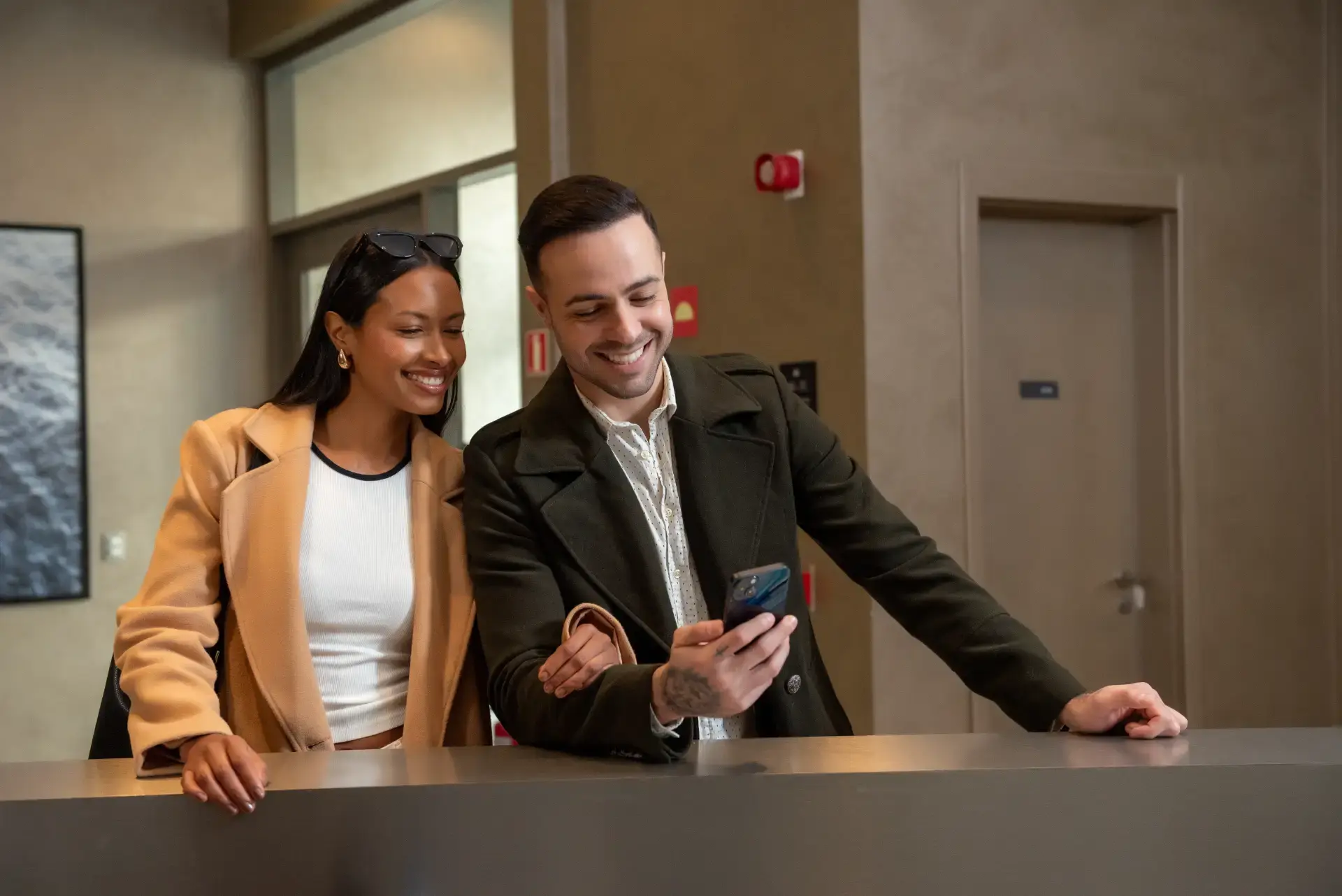 Casal sorridente em um escritório moderno, analisando o celular enquanto conversa à mesa, sugerindo atendimento ou parceria profissional no ambiente corporativo.