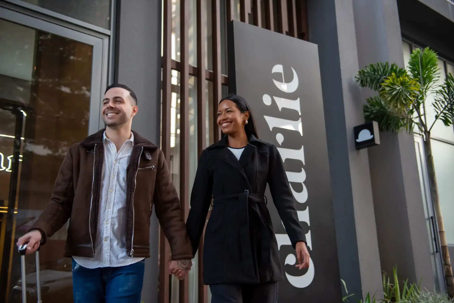 Casal sorridente caminhando de mãos dadas em frente a uma fachada moderna com plantas e porta de vidro, em ambiente externo com luz natural.
