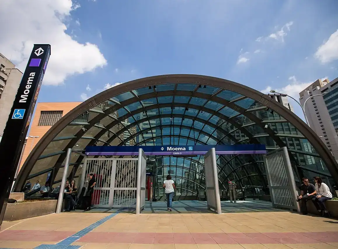 Entrada da estação de metrô Moema em São Paulo, com estrutura de vidro e pessoas ao redor, sob céu azul com algumas nuvens.