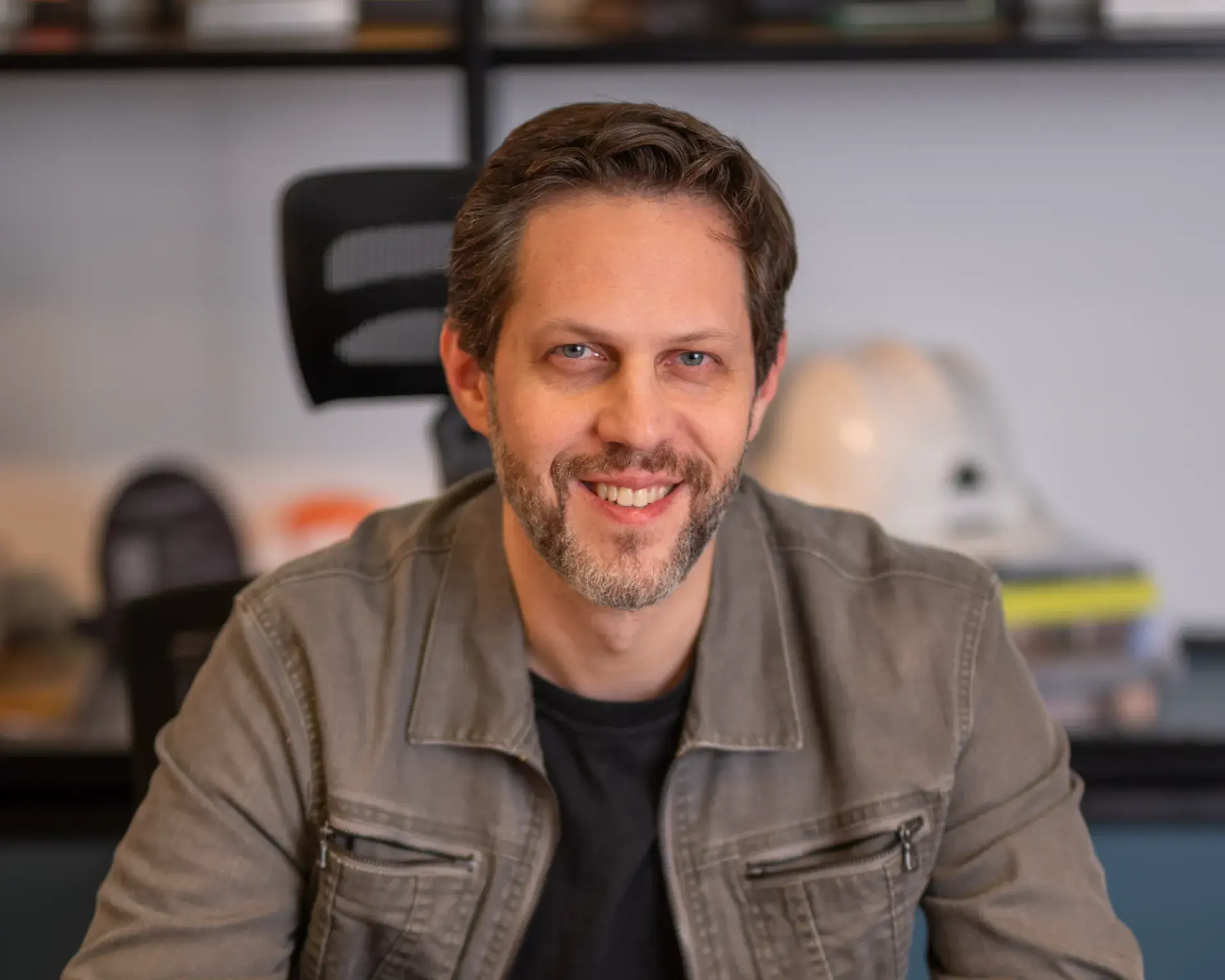 Homem sorrindo em ambiente de escritório, com cabelo castanho e barba curta, vestindo jaqueta cinza sobre camiseta escura, em foto de perfil com fundo desfocado
