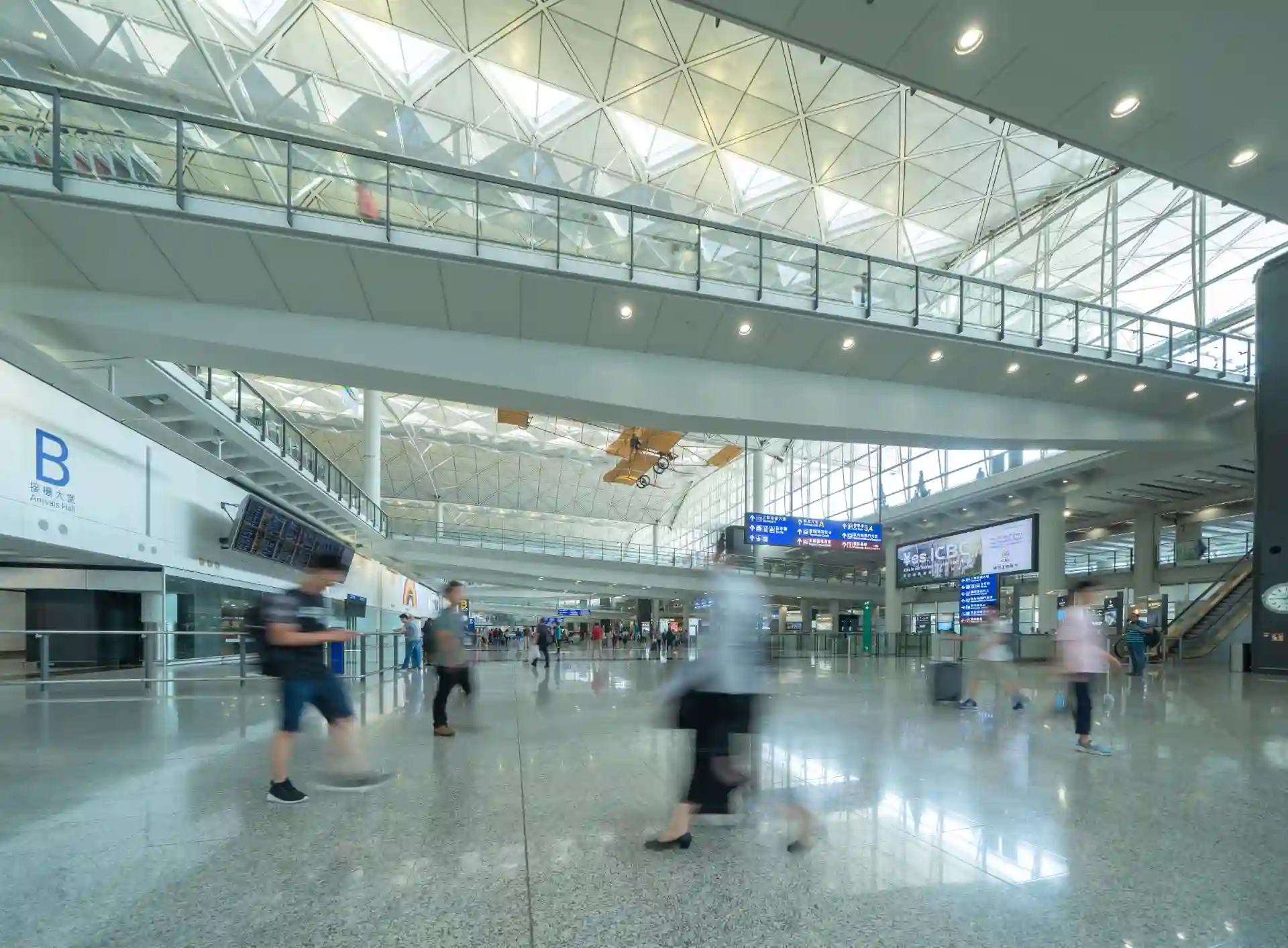 Vista ampla do interior de um aeroporto movimentado com várias pessoas caminhando, um avião decolando no teto e painéis de informações digitais.