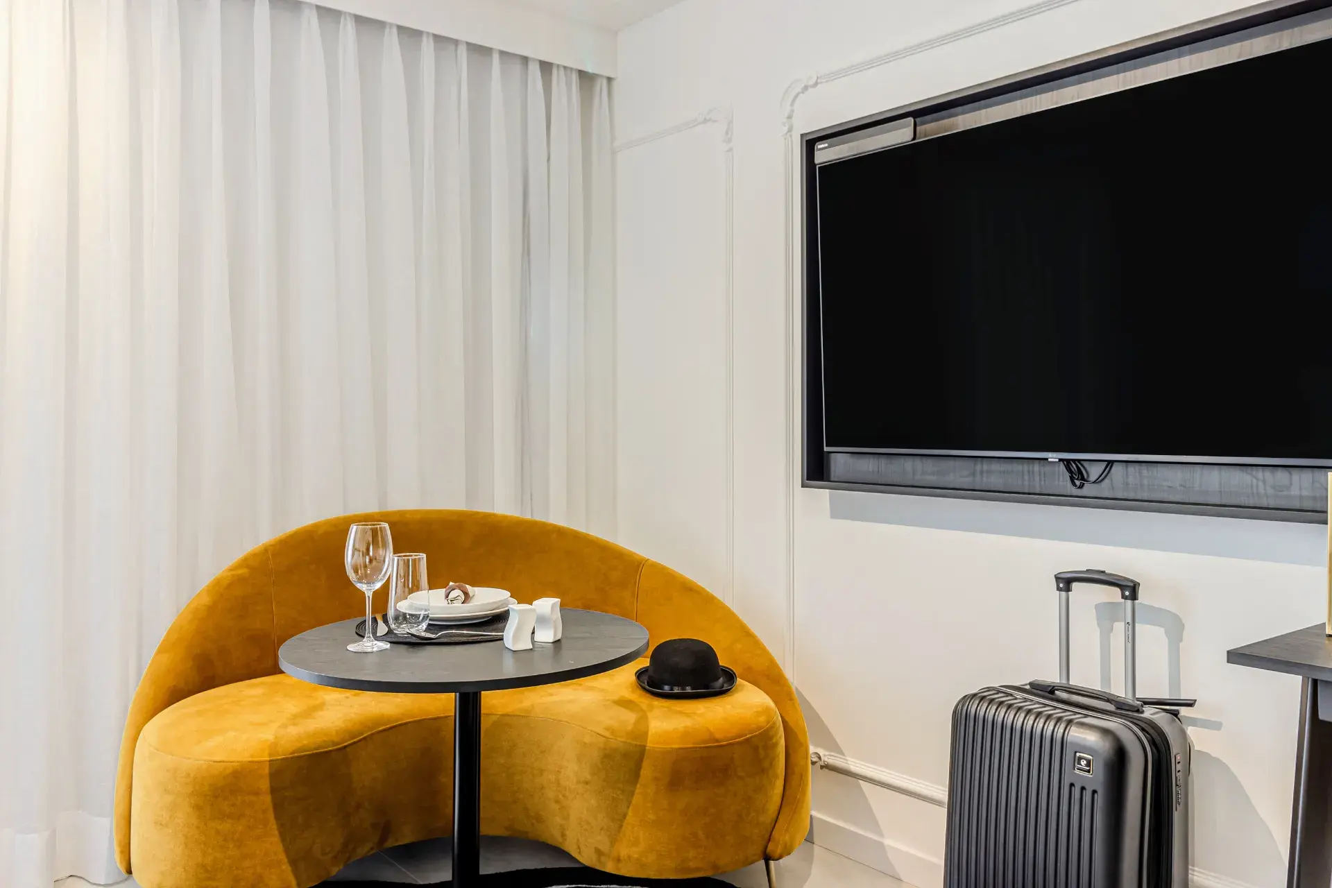 Interior de sala de hotel com poltrona amarela arredondada, mesa redonda com copo e xícara, televisão na parede e mala de viagem de fibra no canto próximo à cortina clara.