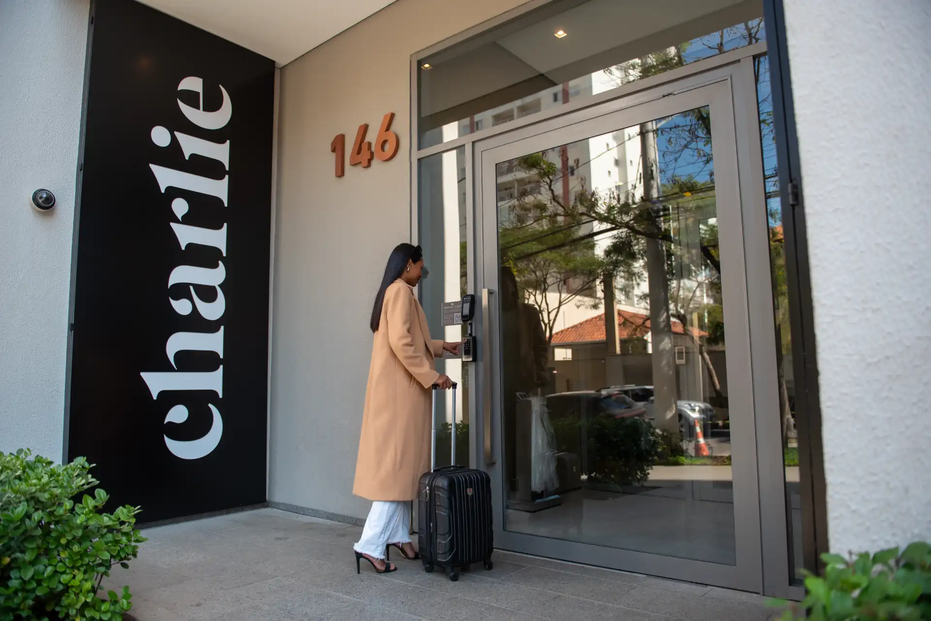Mulher em frente a uma porta de hotel com a placa “Clara Hotel” e número 146, usando um telefone e ao lado de uma mala de viagem preta no saguão externo.