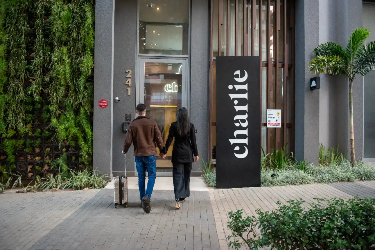 Pessoas caminhando em frente a uma entrada de hotel com placa “Charlie” e número 2411, cercada por plantas e jardim ao lado da fachada.