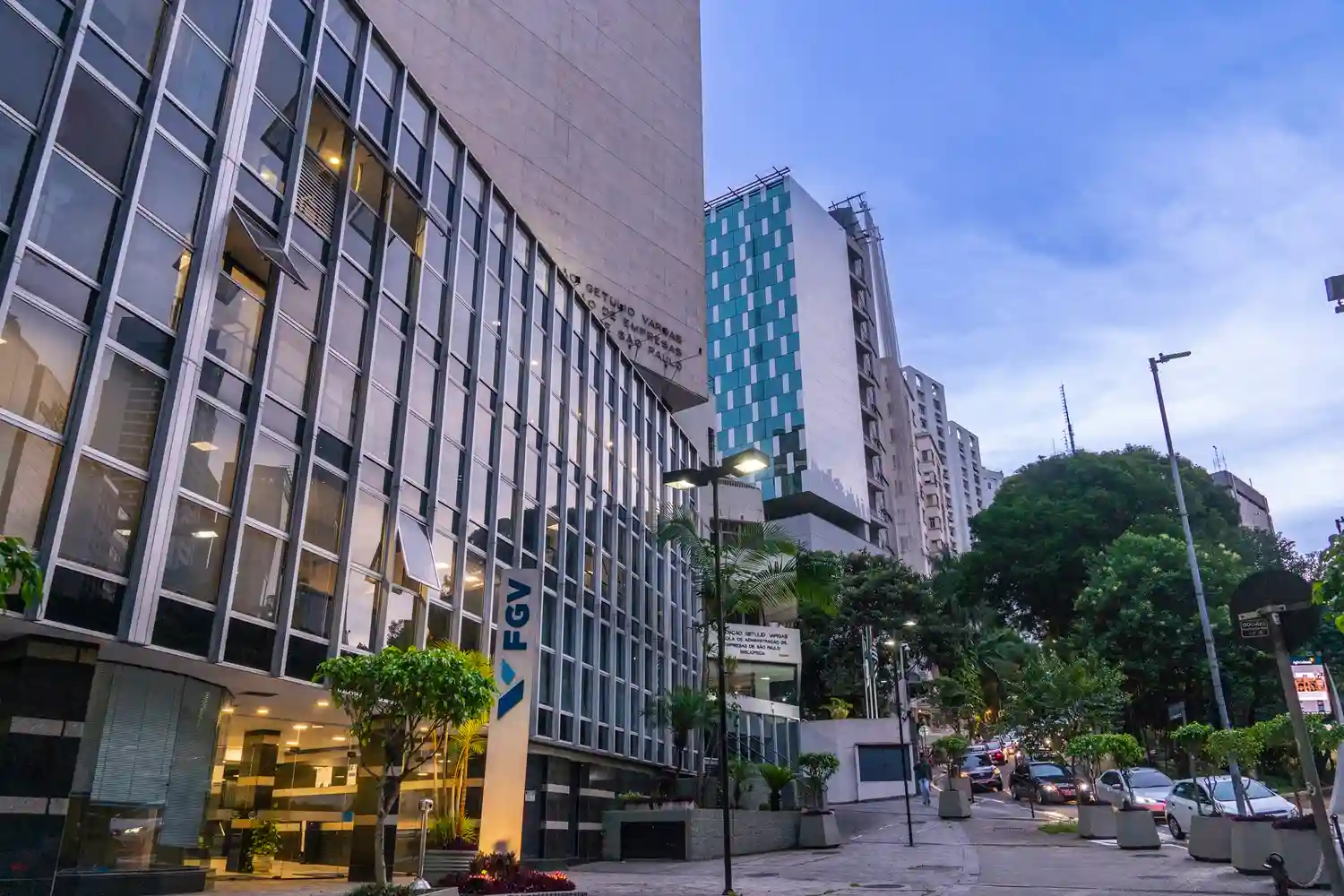 Imagem da fachada de um prédio comercial na cidade, com arquitetura moderna, árvores ao redor, iluminação de rua e veículos estacionados na via.