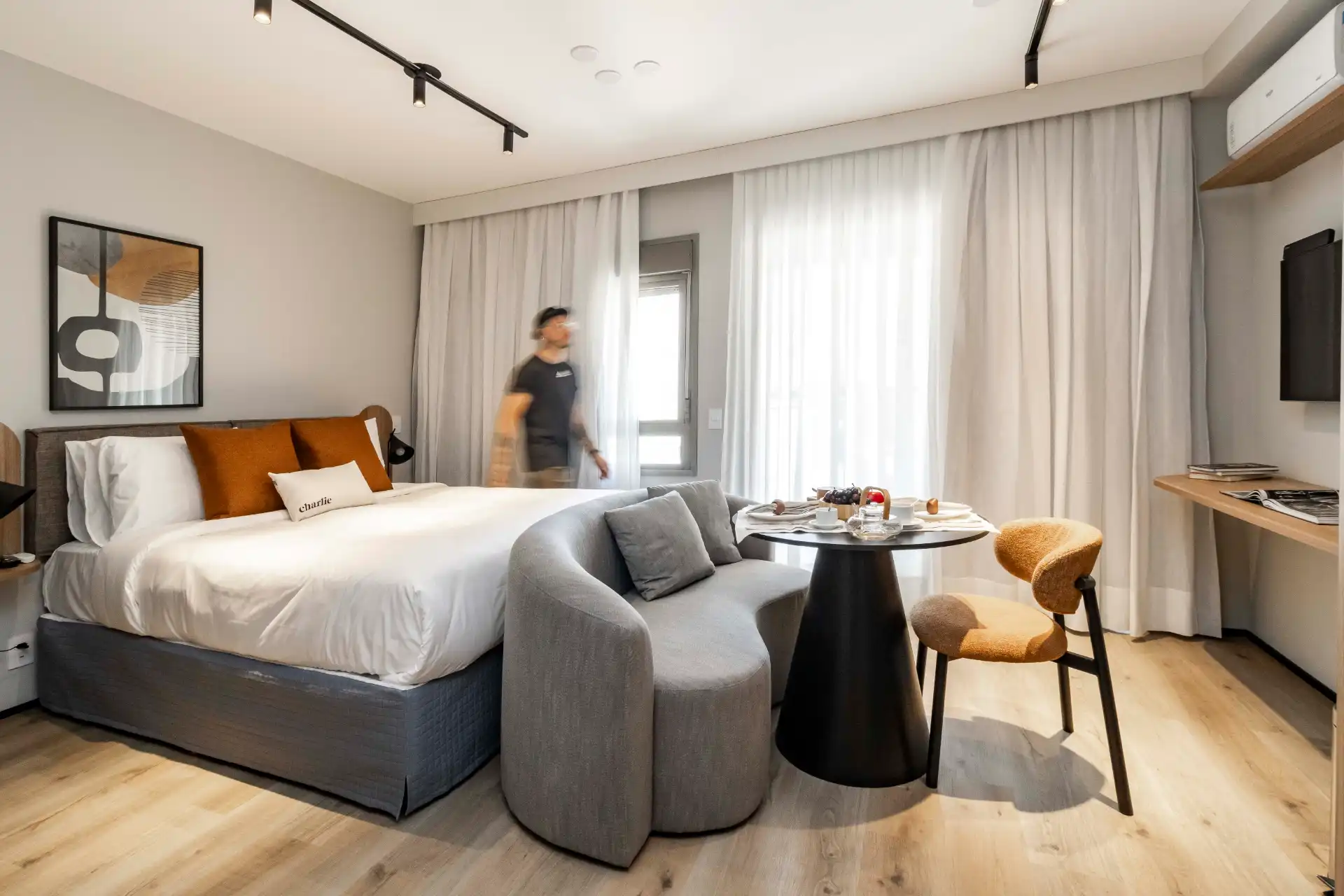 Quarto de hotel moderno com cama de casal, sofá cinza, mesa de jantar, duas cadeiras e cortinas brancas, ambiente bem iluminado.