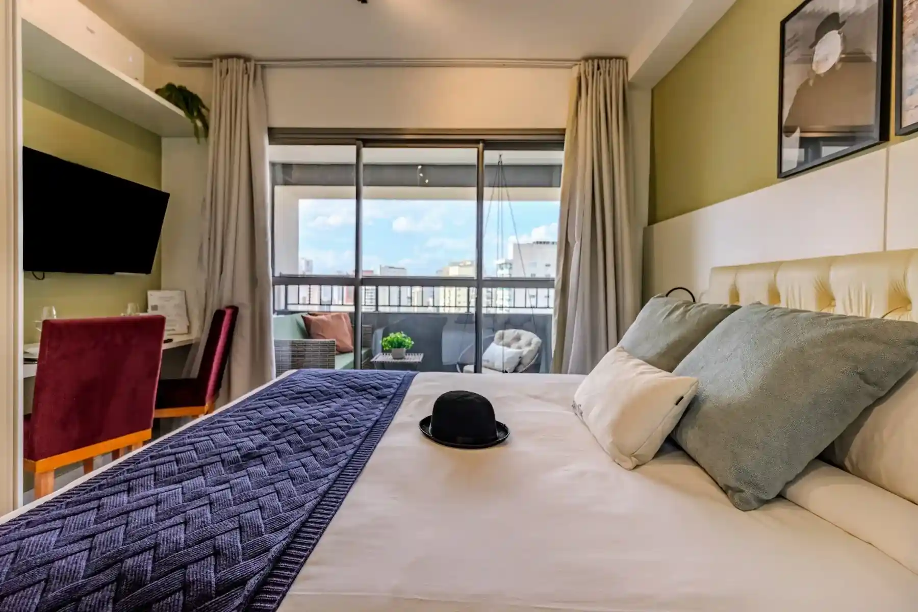 Quarto de hotel moderno com varanda, vista para a cidade, cama com roupas de cama cinza e decorativo preto, televisão na parede, mesas laterais e cortinas claras.