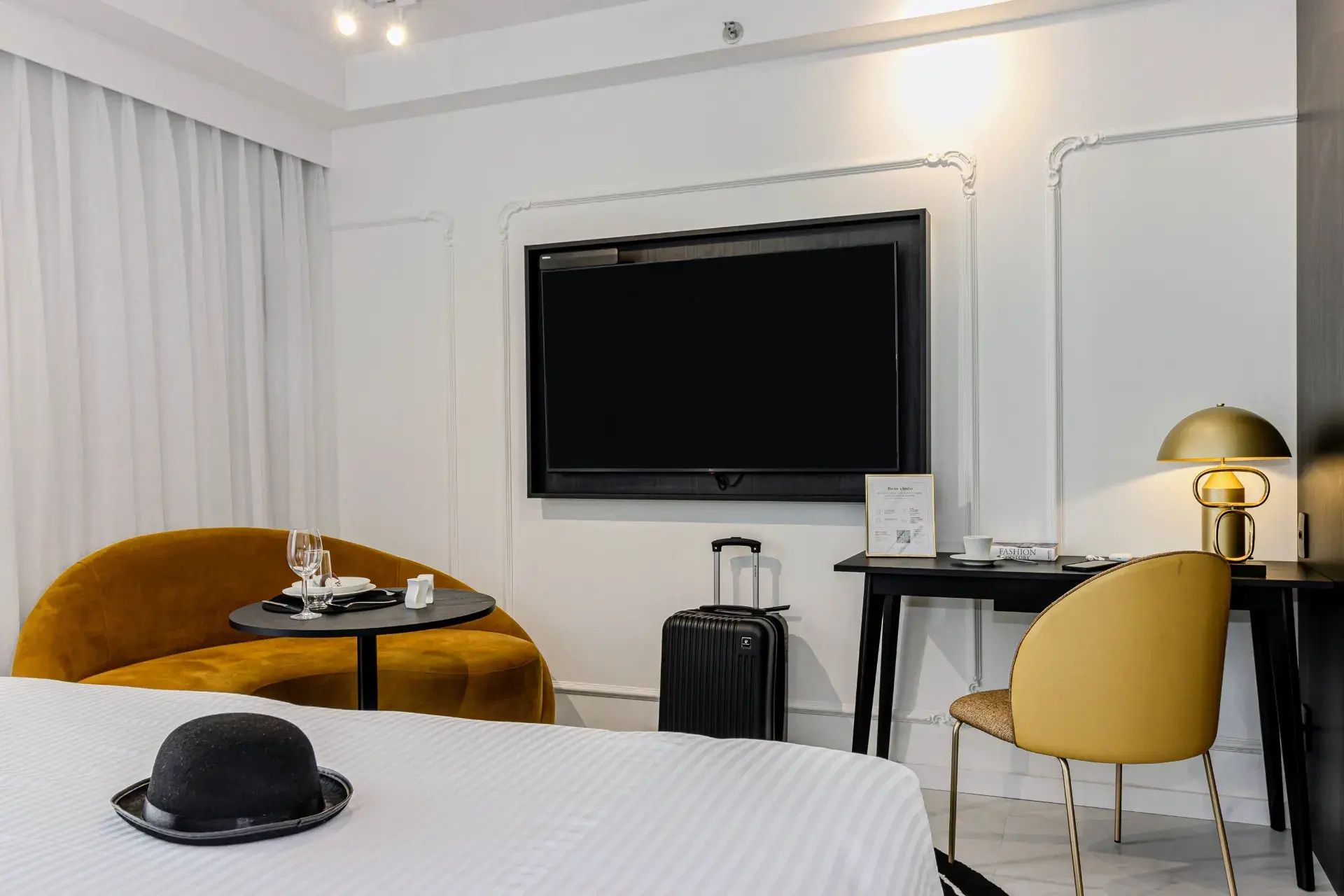 Quarto de hotel moderno com cama clara, poltrona amarela, mesa lateral e TV de parede, iluminação embutida e chaleira/armarinho ao lado