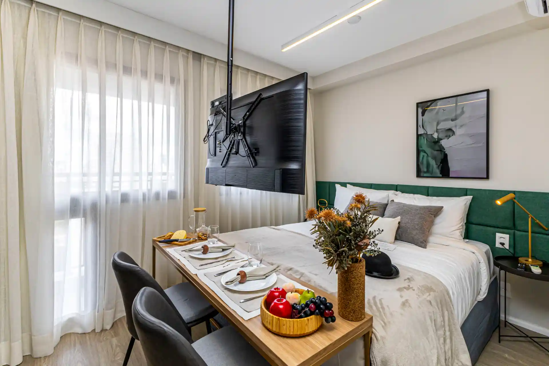 Imagem de um quarto de hotel moderno e bem decorado, com cama confortável, televisão pendurada na parede, mesa de jantar com comida e flores, ideal para viajantes que buscam conforto