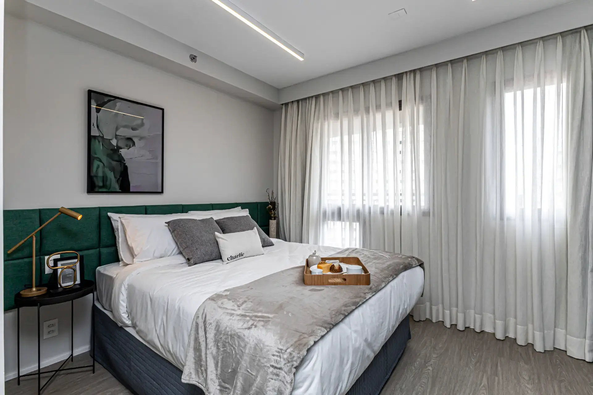 Quarto moderno com cama de casal, roupa de cama branca e cinza, cabeceira verde, mesa lateral com luminária dourada e bandeja com café, além de cortinas claras e luz natural.