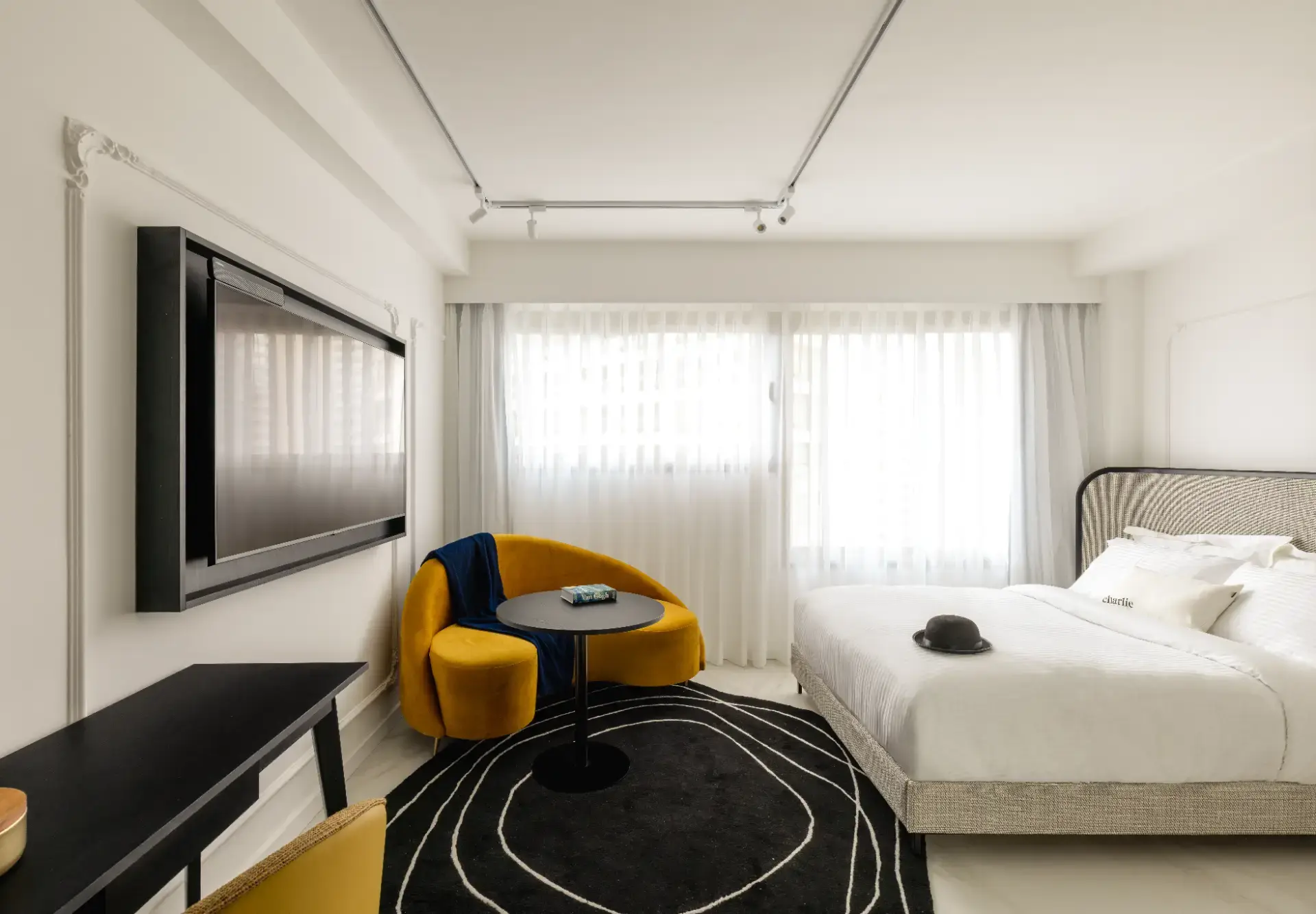 Quarto moderno e iluminado, com cama de casal em base cinza, poltrona redonda amarela, mesa lateral, tapete preto com detalhes claros e cortinas brancas de janela