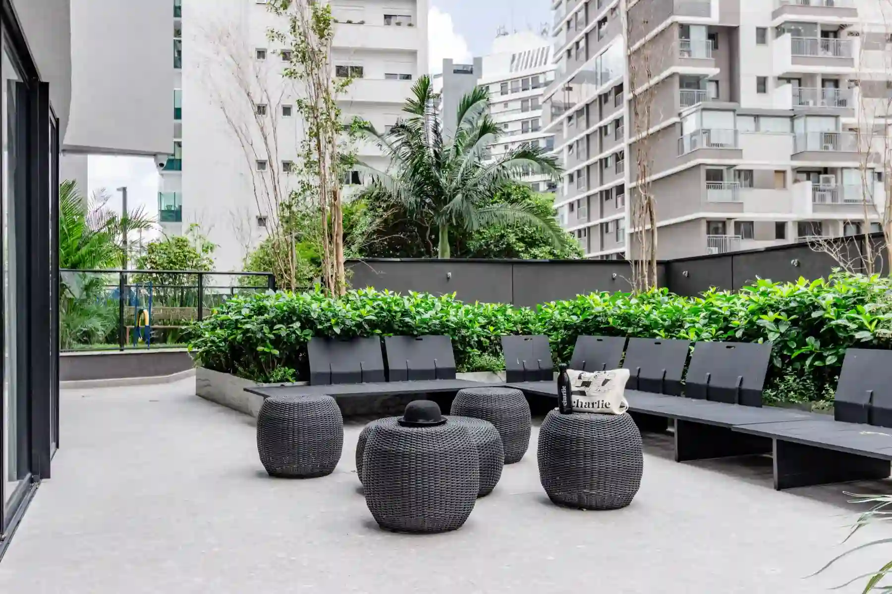 Varanda moderna com móveis pretos de fibra sintética, incluindo cadeiras e pufes, rodeada de vegetação e vista de prédios altos na cidade.
