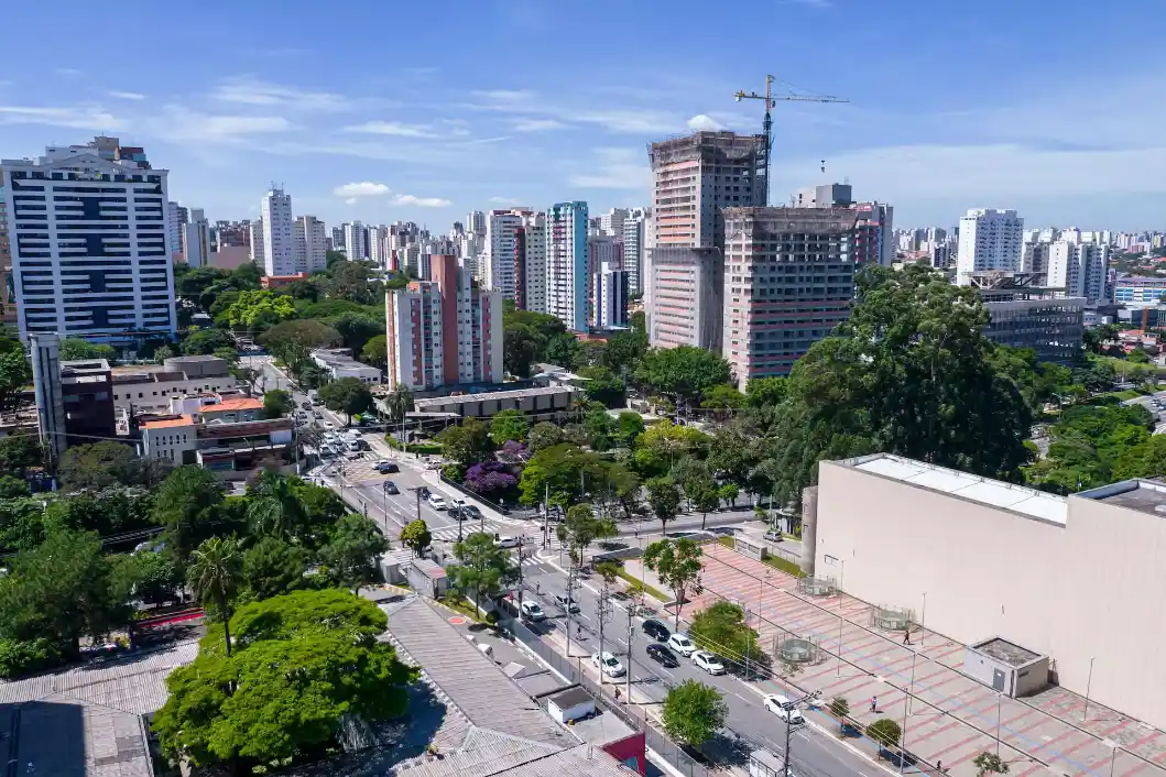 Vista aérea de uma avenida urbana com prédios altos, áreas arborizadas e carros circulando sob céu azul e nuvens, em uma paisagem de cidade em crescimento