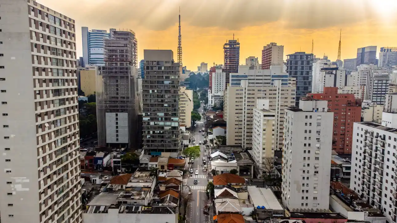 Vista panorâmica de uma avenida em meio a prédios altos e apartamentos, com skyline urbano ao pôr do sol e o céu em tons dourados